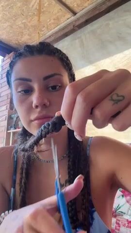 This close-up shot features a young woman with dark, braided hair and a nose ring. She is holding a pair of blue-handled scissors in her right hand, and with them, she appears to be cutting a braid of her own hair. Her left hand is holding the braid steady. She has a tattoo of a small heart on the back of her left hand.

The woman is looking directly at the camera with a neutral expression, though her mouth is slightly pursed, suggesting concentration. She is wearing a blue and white patterned top.

The background suggests an outdoor or semi-outdoor setting. The wall behind her is made of reddish-brown bricks, and there's a wooden beam structure above. A light-colored fabric, possibly a tarp or canopy, is visible in the upper right corner. The lighting suggests it is daytime, likely sunny.

The overall scene is intimate and focused on the woman's action of cutting her hair. There is no visible text in the frame.