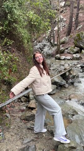 A young woman with long brown hair, wearing a beige puffer jacket, gray sweatpants, and white and blue athletic shoes, stands by a small, rocky stream in a wooded area. She turns to the right, her arm extended, and looks at the camera before turning away and walking across the rocks of the stream. The stream flows rapidly over the rocks, and the surrounding area is lush with greenery and trees. The overall atmosphere is natural and serene.