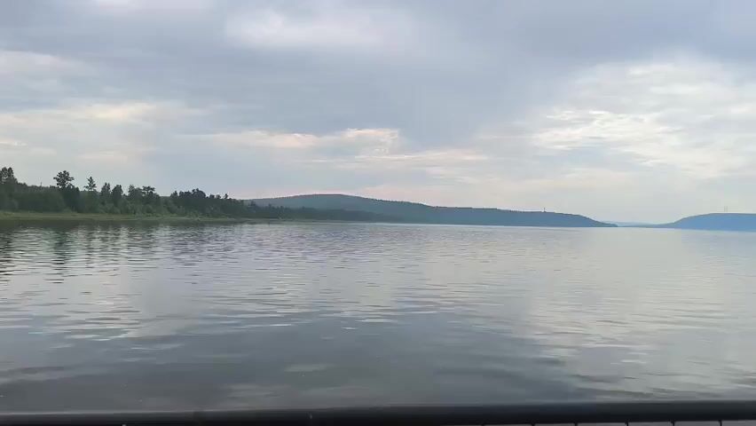 A wide shot of a serene lake under a cloudy sky. The water is calm, with gentle ripples reflecting the overcast heavens. On the left side of the frame, a lush green shoreline is lined with trees, creating a natural border. In the distance, rolling hills stretch across the horizon, their forms softened by the hazy atmosphere. The scene evokes a sense of tranquility and natural beauty.