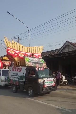 The video shows a parade with two small trucks decorated with flags and banners. The first truck, in the foreground, is dark gray and has a banner on the windshield that reads "TIM CHENNAL AZIZAH." It is carrying a large, elaborate wooden structure decorated with red and white fabric. A person is visible inside the truck's cabin. Behind this truck is a white truck, also decorated with flags and banners, pulling a similar wooden structure. Both trucks are moving forward, suggesting a procession. The environment appears to be a street in a residential area, with houses visible on the right side of the frame and power lines overhead. The lighting suggests it is daytime. The overall scene depicts a festive or celebratory event.
