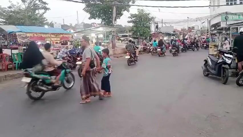 The video shows a street scene with several people and motorcycles. On the left side of the road, there are market stalls with various goods displayed. People are walking and crossing the street. Several motorcycles are seen moving in both directions. One motorcycle is on the left side of the screen, moving away from the camera. Another motorcycle is in the foreground, moving towards the camera. The sky appears overcast. The scene is bustling with activity, suggesting a public area with commercial activity.