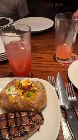 The video captures various moments of people dining in a lively restaurant setting in Schaumburg, United States of America, likely during the evening. Multiple tables are visible, laden with a variety of food and drinks.

The food items shown include a plate with ribs, macaroni and cheese, and a loaded baked potato. Another plate features grilled corn on the cob, a steak or filet, and fried rice. Salads, bread, and other side dishes are also seen. Drinks vary from red cocktails and margaritas to soft drinks.

Several individuals are present, some seen talking, smiling, and posing while enjoying their meals. One person holds up a large piece of meat, possibly ribs. The atmosphere appears casual and social, with people gathered around wooden tables and booth seating. A cocktail shaker and a seasoning bottle are visible on one of the tables.