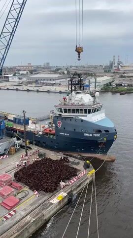 A wide shot captures a port scene with a large ship, the "SHAW TIDE," docked alongside a pier. A massive blue crane looms on the left, its arm extended over the water. A smaller crane, attached to a barge, is visible further in the distance. The "SHAW TIDE" is dark blue with a white superstructure and red accents. Containers are stacked on its deck. In the foreground, a large pile of dark, irregular objects, possibly scrap metal or debris, sits on the pier. A person wearing a yellow hard hat is visible on the pier near the ship, appearing to be securing ropes. The sky is overcast. The water is calm and reflects the sky and structures. The background shows industrial buildings and structures.