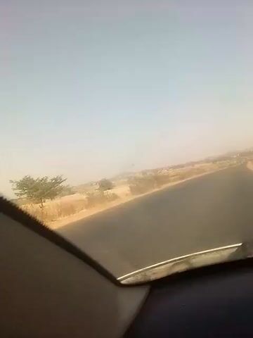 The video depicts a view from inside a car driving on a paved road. The sky is a pale, hazy blue, suggesting a clear day. The landscape on either side of the road appears dry and arid, with sparse vegetation and low-lying buildings visible in the distance. The camera angle is low, positioned at the dashboard level, showing the edge of the dashboard and the windshield. The car seems to be moving at a steady speed, as indicated by the blurring of the peripheral scenery. The overall atmosphere suggests a remote or rural location.