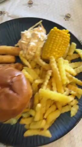 A medium shot displays a plate of food. The plate is dark blue and has vertical lines across it.  A hamburger with a brown bun sits on the left side of the plate. The burger appears to have lettuce, onion, and cheese inside. Next to the burger is a pile of crinkle-cut french fries.  Above the fries is a cob of corn on the cob and a scoop of coleslaw.  To the left of the burger are what appear to be breaded mozzarella sticks. The food sits on a table with a floral pattern. An unseen person is recording the meal. Voices can be heard in the background, one speaking the word "green."