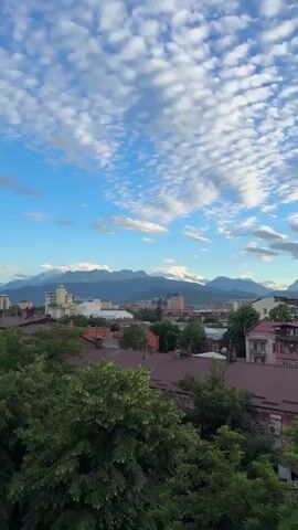 The video displays a cityscape with mountains in the background. The sky is blue with scattered white clouds. In the foreground, trees with green leaves are visible. Several buildings of varying heights and architectural styles are present in the city, some with reddish-brown roofs. In the distance, a range of majestic mountains, some capped with snow, rises against the horizon. A Ferris wheel-like structure is partially visible on one of the mountains. The overall impression is a scenic view of a city nestled near a natural mountain landscape.