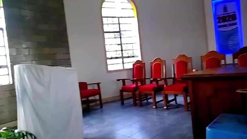 The video shows an interior of a church. The first scene shows a view of a wall with a window, four red chairs, a brown podium, and a blue crate. There is a poster on the wall behind the podium. The next shot is a closer view of the windows on the side of the church, with some windows having colored stained glass. The camera then pans to a wide shot of the interior showing rows of wooden pews and additional windows. The camera then focuses again on a row of arched windows with what appears to be a wall of gray bricks.