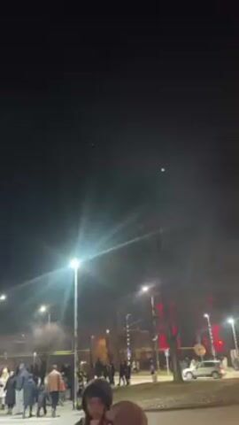 The video shows a crowd of people standing on a street at night. Streetlights illuminate the area, creating bright points of light with starburst effects. Smoke drifts through the air, partially obscuring the background. Buildings with red lighting can be seen in the distance. Some cars are parked along the street. A couple of people stand in the foreground, partially blocking the view. The overall atmosphere suggests a public gathering or event.