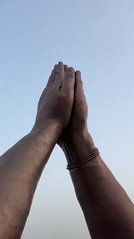The video features a large statue of two hands in a praying position, with a bracelet on one wrist. The statue is made of bronze or a similar material, and the sky is a clear, light blue. In the background, there is a bridge over a river and some people are visible. The camera pans slightly to show more of the surrounding area and people before returning to focus on the hands.