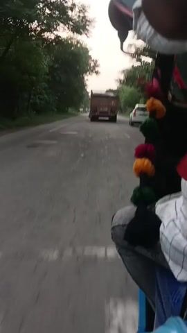 The video is taken from the perspective of a passenger in a vehicle, possibly an auto-rickshaw, traveling on a road. The road is asphalt and appears to be in disrepair with visible patches and cracks. Lush green trees line both sides of the road, with a power line pole visible on the left side. In the distance, a large brown truck is traveling in the same direction, followed by a white car. A white cow is standing near the side of the road on the left. The sky is overcast, suggesting it may be late afternoon or early morning. Colorful tassels or pom-poms are hanging from the vehicle's interior on the right side of the frame. The passenger's leg and arm are visible in the foreground.