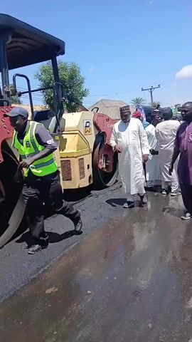 A group of people are walking down a street past a yellow road roller. Some of the people are holding up posters with pictures of men on them. The street appears to be under construction, with asphalt visible. The sky is blue with some clouds.