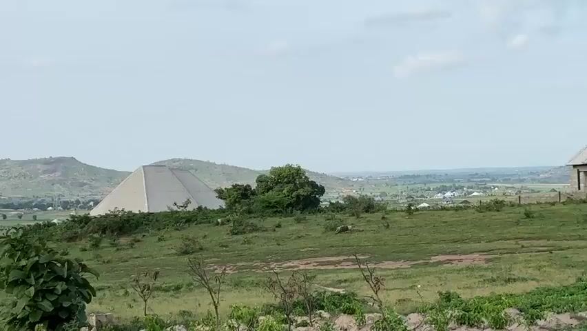 The video pans across a rural landscape. In the foreground, there are some unfinished brick structures and green vegetation. In the mid-ground, a large, pyramid-shaped building with a white roof stands out amongst the green hills. Several other smaller, unfinished buildings are scattered throughout the landscape. In the background, rolling hills and a distant town can be seen under a cloudy sky. The overall impression is one of development in a natural setting.