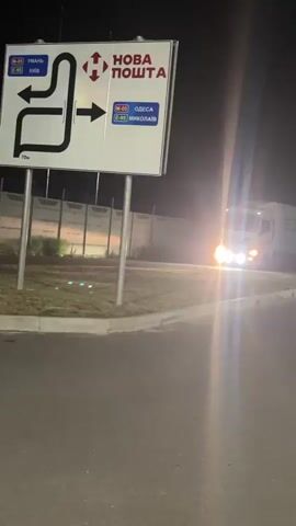 A white semi-truck is turning right at night in front of a large sign that reads "Nova Poshta" with directional arrows. The truck's headlights are on and are illuminating the road and surrounding area. The sign has directions for Kyiv, Odesa, and Mykolaiv. There is a barrier gate and a yield sign visible near the truck. The ground appears to be asphalt or concrete. The background shows a concrete wall and some lights in the distance.