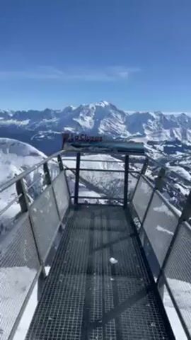 The video shows a metal viewing platform with a sign that reads "La Clusaz" with a landscape image on it. Behind the platform is a mountain range covered in snow. The sky is blue with some clouds. The camera moves slightly toward the sign. It is windy.