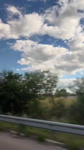 The video shows a view from a moving vehicle, likely a car, looking out at a rural landscape. The scene is dominated by a bright blue sky with fluffy white clouds. The ground below consists of a field of dry grass and scattered trees and bushes. In the distance, a small cluster of houses can be seen. A metal guardrail runs along the bottom of the frame, indicating the perspective is from a road. The motion of the vehicle causes the landscape to blur, especially the trees and bushes. The sun appears to be shining, casting a bright light on the scene.