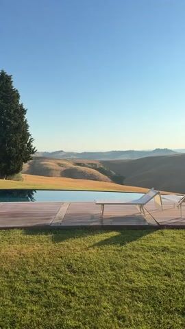 The video pans across a serene landscape, showcasing a swimming pool on a wooden deck. Two lounge chairs are placed on the deck, facing the rolling hills in the distance. The sun is shining brightly in a clear blue sky, casting a warm glow over the scene. A tall, slender cypress tree stands prominently on the right side of the frame, adding a touch of verticality to the predominantly horizontal landscape. The foreground is a lush green lawn, creating a peaceful and inviting atmosphere. The overall impression is one of tranquility and natural beauty.