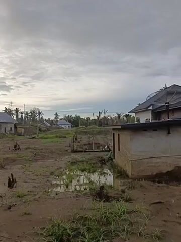 The video shows an exterior view of a village with several houses. The ground is muddy and uneven, with some puddles of water. There are tree stumps scattered throughout the area. The sky is cloudy, and the overall lighting suggests it is either early morning or late afternoon. Several houses are visible, some appearing to be under construction or unfinished. In the foreground, there is a small, partially built structure on the right. In the background, more houses and palm trees are visible. The scene appears to be a rural or semi-rural settlement. No people are clearly visible in the video.