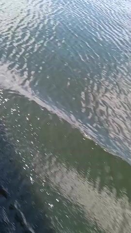 The video shows a close-up, angled shot of water. The water's surface is rippled, creating patterns of light and shadow. The water is darker in the foreground and transitions to a lighter shade towards the background. There are some reflections visible on the water's surface. The shot appears to be taken from a moving perspective, possibly from a boat or by someone walking near the water.