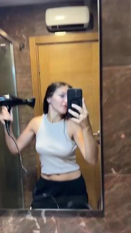 A woman with long dark hair stands in a bathroom, taking a selfie in a mirror. She is wearing a beige tank top and black sweatpants. She holds a black hairdryer in her right hand, pointing it towards her hair. Her left hand holds a smartphone, angled to capture her reflection. She is making a playful face, sticking her tongue out slightly. The bathroom features marble walls, a wooden door, a glass shower enclosure, and a white air conditioner unit mounted on the wall above the door. The lighting is bright, illuminating the scene.