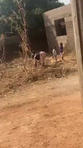The scene depicts a rural environment, possibly a village. A person is bent over, working in a dry, grassy area with scattered debris. To the right of the person, two children stand near a partially constructed brick building. The ground is a red-brown dirt. The background consists of trees and the unfinished building, suggesting a developing or impoverished area. The light indicates it is daytime.