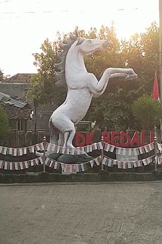 A white horse statue is in the foreground of the video. The statue is facing left, with its head up and legs in the air. The horse is decorated with a red and white ribbon. Behind the horse is a sign with red letters that say "DK BEDALI". There are trees and houses in the background. The video is filmed from a low angle.