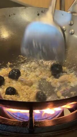 The video shows a cooking process using a wok over a high-flame gas burner. The wok contains a mixture of ingredients, including small pieces of what appears to be chopped tuna, minced onions, and several whole, dark-colored olives. A shiny, silver spatula is used to stir and toss the ingredients within the wok, ensuring even cooking. The cooking surface inside the wok is shiny and oily, reflecting the light. The gas burner underneath the wok produces a bright blue flame with orange tips, indicating intense heat. The wok itself is made of dark metal and shows signs of previous use. The backdrop is blurred, suggesting an indoor kitchen or food preparation area.