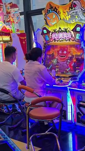 A man and a woman are sitting on chairs in front of a colorful arcade game. The man is on the left, wearing a white shirt, and the woman is on the right, wearing a pink jacket and white shorts. They are both facing the game, which has a Halloween theme with a pumpkin and a monster on the screen. The woman is holding a joystick and appears to be playing the game. The arcade is brightly lit with blue and pink neon lights. Other arcade machines are visible in the background.