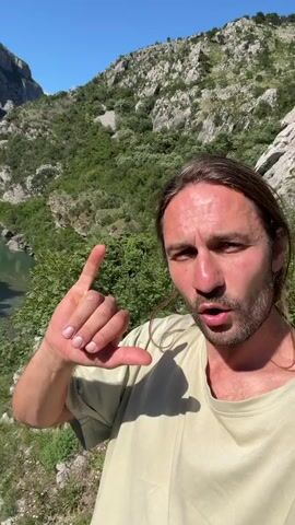 A man with long dark hair and a beard, wearing a light-colored t-shirt, films himself in a selfie style. He holds his left hand up with his thumb and pinky extended, then points downward with his thumb towards a metal suspension bridge. Below the bridge, a turquoise river flows through a deep gorge. Two individuals are walking on the bridge; one in white clothing is further away, and another in dark clothing is closer. The background features steep, rocky cliffs covered with green vegetation under a clear blue sky.