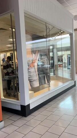 A wide shot of a shopping mall interior shows a storefront with large glass windows displaying mannequins in jeans. To the right, the mall corridor stretches out, with polished tile floors reflecting overhead lighting. Several other storefronts and structural pillars are visible in the distance, along with some seating areas and potted plants. The scene is quiet and appears to be sparsely populated.