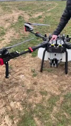 A man in a black jacket stands in a field next to a large drone. The drone is black with red accents and has six propellers. It has a white tank mounted underneath. The man is pressing buttons on top of the drone. The field is mostly brown dirt with some green patches of grass. In the background, there is a row of trees and a gray sky.