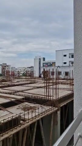 The video shows a construction site with a cloudy sky. In the foreground, there are concrete slabs and rebar forming the framework of a building. In the middle ground, a man is walking across the construction site. In the background, there are several buildings, some of which appear to be residential apartments. The overall scene depicts ongoing construction work in an urban environment.