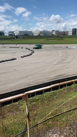 A green sedan is drifting around a track marked with tires. The track is paved and appears to be outdoors. In the background, there are buildings and a tower. A person is standing on the track near the car in one frame. There is a wire fence in the foreground, with grass and dirt behind it. The sky is cloudy.