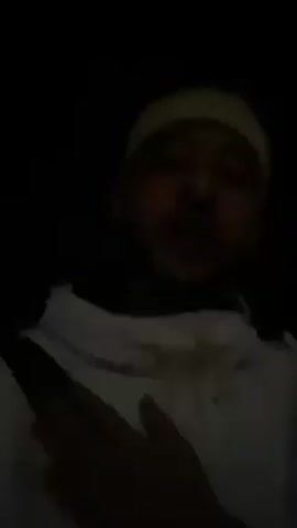 The video shows a man in a white jacket and off-white beanie. He is wearing a seatbelt. He is rapping while looking at the camera, making hand gestures. The background is dark.