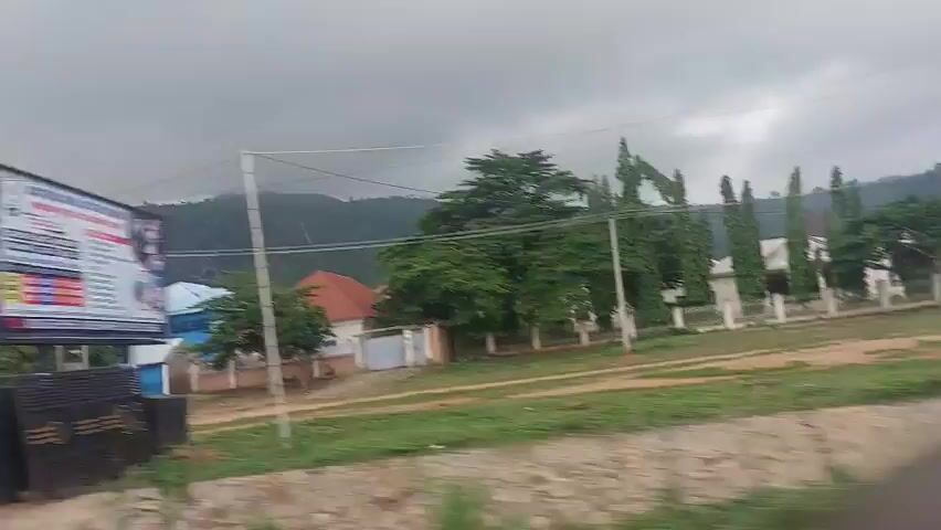 The video captures a street view with buildings and a mountain in the background. The sky is overcast. Several multi-story buildings, some painted blue and others with red roofs, are visible. There are trees scattered throughout the scene. A utility pole with power lines runs across the frame. In the foreground, there is a brown and beige three-wheeled vehicle with people inside, moving from left to right. Several cars are parked along the street. Construction materials, including metal pipes, are present on the left side of the frame. The ground is a mix of dirt and grass.