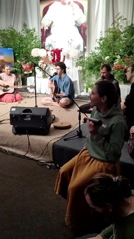 A group of people are gathered in a room, possibly a temple or community hall, for a musical performance. In the center, a young man with glasses and dark curly hair is playing an acoustic guitar and singing into a microphone. He is seated cross-legged on a raised platform covered with a brown rug. To his left, a woman with blonde hair tied back is also playing an acoustic guitar and singing. She is wearing a pink top and a pink skirt. To the right of the guitarist, a woman in a green hoodie and a long orange skirt is holding a microphone and appears to be singing or speaking. Behind her, a man with short dark hair is also holding a microphone. In the background, a large portrait of a person in white robes is displayed, adorned with floral arrangements. The stage is decorated with green foliage and red and white flowers. A black speaker is placed on the platform in front of the guitarist. Several other people are visible in the foreground, some seated and some partially obscured. The lighting is warm and ambient.