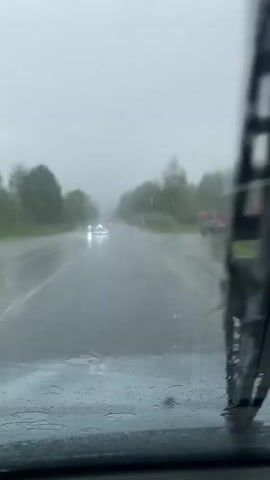 The video shows a view from inside a car on a rainy day, looking through the windshield. Raindrops are visible on the glass, and the windshield wipers are intermittently clearing the view. The road ahead is wet and appears somewhat foggy. Several cars are driving in the distance, their headlights illuminated. Trees and foliage line the sides of the road, and the overall environment suggests an overcast and wet outdoor setting.