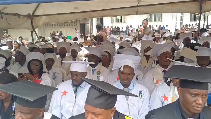 A large crowd of people wearing white graduation gowns and mortarboard caps sit under a temporary tent structure. Many of the caps are black, while others are white. A man standing to the right holds a cell phone in his hand as if he is taking a photo or video. Behind him, other people are standing and sitting, possibly watching the graduation ceremony. The scene appears to be taking place outdoors on a sunny day, with a building visible in the background. There are a number of students sitting in rows. Some are looking down or towards the ground. In the foreground, a woman is wearing earrings and seated. The video appears to be taken from an elevated position. A large sign partially visible reads "ECOLE DE SANTE PUBLIQUE." The scene is crowded and filled with graduates and spectators.