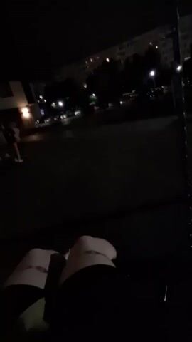 The video begins with a first-person perspective of someone's legs wearing ripped jeans and white sneakers. The camera is low, pointing upwards. The setting appears to be outdoors at night, with buildings and streetlights visible in the background. The camera then pans upwards to show a dark sky with some lights from buildings. The view shifts again, showing a group of people in the distance, some of whom are moving. One person in the distance is wearing a white top. The camera continues to pan and tilt, showing more of the night environment, including trees. The video ends with a brief shot of a dark sky. The overall scene suggests a casual outdoor gathering or activity at night.