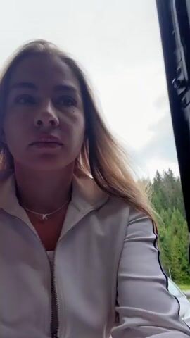 A woman with long blonde hair is in the back of a vehicle. She is looking down at the camera and appears to be speaking. She is wearing a white zip-up jacket over a white shirt. She has a necklace with a "K" pendant. The interior of the vehicle is visible, including the headliner and a light fixture. Outside the vehicle, greenery is visible. The woman then looks up and smiles broadly at the camera as she moves.