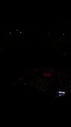 The video shows a dark, outdoor scene at night. A large crowd is present, with many people holding up illuminated objects, possibly phones or lights, which appear as red and white dots. The location is unclear, but it appears to be an open space with sparse overhead lighting. A person in the foreground is holding a mobile phone, with its screen visible. There is some ambient noise in the background, including music and cheers, suggesting the event might be a concert or performance.