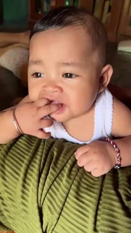 A baby is lying on its stomach on a green fabric, facing the camera. The baby is wearing a white tank top and has bracelets on both wrists. The baby is chewing on its fingers. A person's hand enters the frame and gestures towards the baby. The baby looks at the hand and then back at the camera. The background is blurry and appears to be an indoor setting with furniture and other objects.