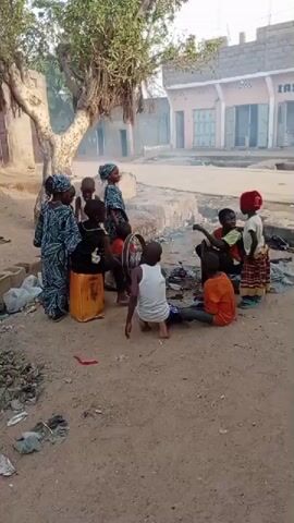 A group of approximately 10 children and women are gathered in a sandy area. Some are sitting on the ground, while others are standing around a yellow container. There is a bicycle wheel present, and some of the individuals seem to be interacting with it. In the background, there are buildings and a tree, suggesting an outdoor village or residential setting. A figure in dark clothing is walking on the road in the background. There is some scattered trash on the ground and some visible smoke in the air.