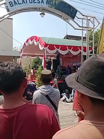 A low-angle, medium shot shows a group of people standing outside, facing away from the camera. In the foreground, the backs of three individuals are visible: one wearing a maroon t-shirt, another a grey hoodie and a peaced cap, and the third wearing a black hat. Behind them, an archway with the text "BALAI DESA NG..." is visible. Beyond the archway, a stage is set up with a red and white tent canopy. A flag of Indonesia is displayed on the stage. Several people are on the stage and in front of it, with some audience members sitting on the ground. The environment appears to be outdoors with bright daylight.