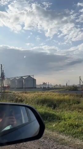 The video shows a view from the side mirror of a car. A person's reflection is visible in the mirror, holding a phone. The car is moving on a gravel road. In the background, there is a field of dry grass and tall trees. Beyond the field, there are industrial buildings, including several large grain silos and a smaller building. A power line with a tall pole is also visible. The sky is partly cloudy with the sun shining through. There are a few cars parked near the industrial buildings. Dust is seen being kicked up near the silos, suggesting some activity.