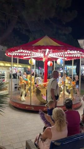 This is a vertical, slightly shaky, nighttime video of a small carousel for children. The carousel is red and yellow, with lights around the edge of the canopy and hanging striped balls. Several children are riding on the carousel horses, which are yellow and white with gold accents. An adult man is operating the carousel. In the foreground, two women are sitting on a bench facing the carousel. One woman is wearing a pink top and holding a purple fan, and the other is wearing a dark pink top. The background shows a street scene with lights and several shops. The carousel is located outdoors on a paved area.