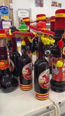 The video displays a collection of souvenir bottles filled with dark red liquid, presumably Sangria. The bottles are shaped like human torsos with elongated necks and two bulbous sections near the base, resembling legs. Each bottle is adorned with a novelty Spanish hat, featuring vibrant colors like red, yellow, and green, and some have dangling decorative elements. The labels on the bottles indicate "Sangria," "Corrida," and "Souvenir." The bottles are arranged on a white shelf in what appears to be a store setting, with other products and shelving visible in the blurred background. The lighting is bright, suggesting an indoor environment. There are no people visible in the video.