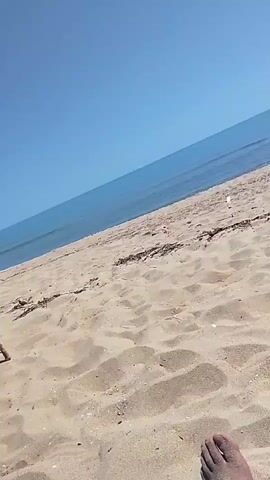 The video shows a sandy beach on a sunny day. The camera is positioned low, near the sand, and focuses on a person's bare foot in the foreground. The sea is visible in the background with gentle waves and a clear blue sky above. There are some pieces of driftwood scattered on the sand near the water's edge. The overall scene is peaceful and depicts a relaxing day at the beach.