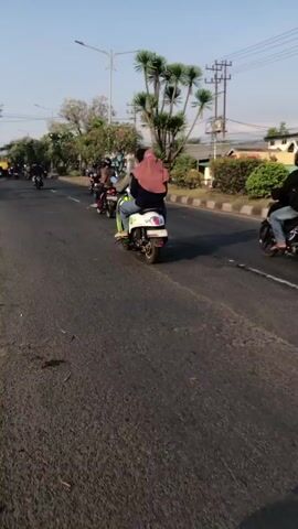 The video shows two motorcycles on a paved road. The first motorcycle, on the left, carries two people, one driving and one passenger in the back. The passenger is wearing a pink headscarf. The second motorcycle, on the right, also carries two people, both wearing helmets. The driver of the second motorcycle is wearing a black jacket and a dark-colored helmet. The motorcycles are moving away from the camera. The environment appears to be a city street with trees, buildings, and utility poles visible in the background. The road is asphalt and appears to be in good condition. The sky is clear and blue, suggesting a sunny day.