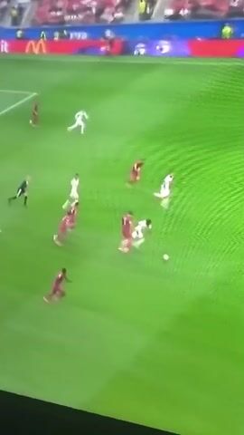 This is a zoomed-out, shaky, overhead video clip of a soccer match. The field is green, and there are white lines marking the boundaries and center circle. Several players in white uniforms and several players in maroon uniforms are on the field. One player in a white uniform is dribbling a soccer ball toward the right side of the frame. Several players in maroon uniforms are attempting to defend him. One player in a white uniform is on the ground near the center circle. The stands are visible in the background and are filled with spectators. Advertisements are visible on the stands.