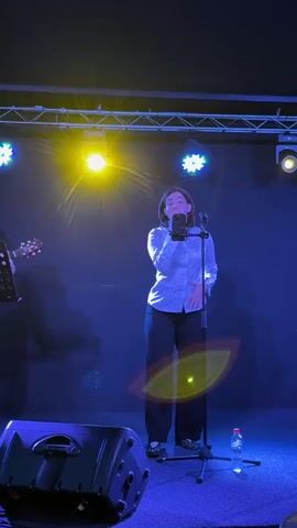 A woman is standing on a stage, singing into a microphone. She is wearing a white, patterned button-down shirt and black pants. Behind her, there are blue and yellow stage lights. To the left of the frame, a guitar is visible. To the right of the frame, a bottle of water is on the floor. The woman is gesturing with her hands as she sings.